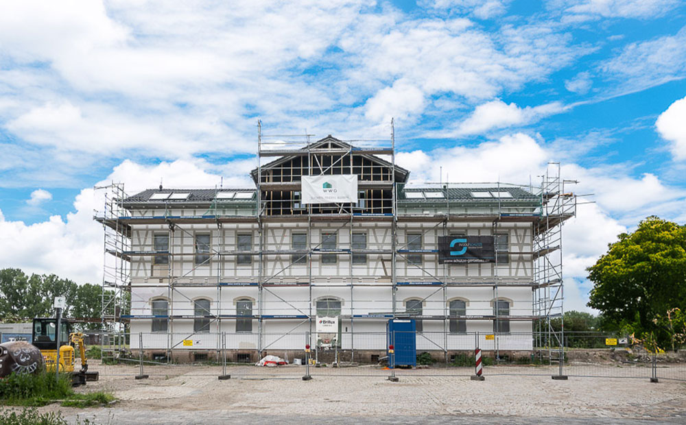 Ein Moment von großer Bedeutung für Kölleda – WWG Kölleda und Stiftung Finneck unterzeichnen Mietvertrag zum Bahnhof 1 Der Bahnhof Kölleda ist vollständig von Baugerüsten eingerahmt und befindet sich in der Sanierungsphase. Das historische Gebäude mit heller Fassade ist mittig im Bild zu sehen. Auf dem Gerüst hängt ein Banner mit dem Logo der WWG Wohnungswirtschaft GmbH. Vor dem Gebäude stehen eine mobile Toilette und ein kleiner Bagger. Der Himmel ist blau mit weißen Wolken, im Hintergrund sind Bäume zu sehen.