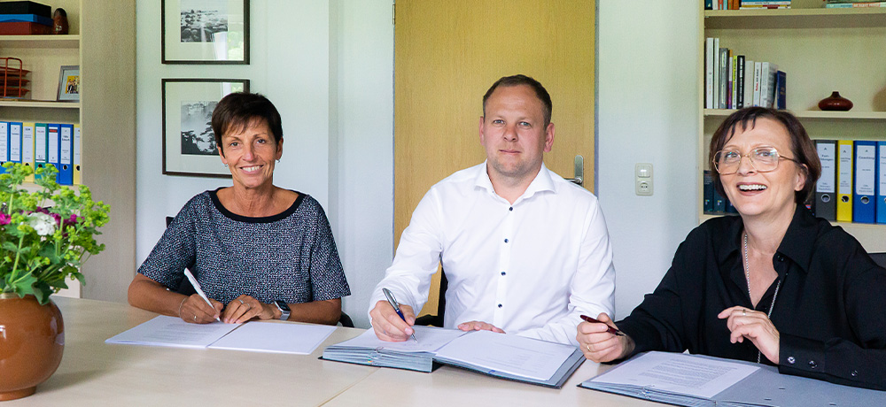 Drei Personen sitzen an einem Tisch und unterzeichnen Dokumente. Von links nach rechts: Silke Kösling (Stiftung Finneck), Kai Vollrath und Christina Otto (WWG Wohnungswirtschaft GmbH). Alle lächeln in die Kamera. Im Hintergrund sind ein Regal mit Ordnern, Bücher und Bilder an der Wand zu sehen. Ein Blumenstrauß in einer braunen Vase steht auf dem Tisch.