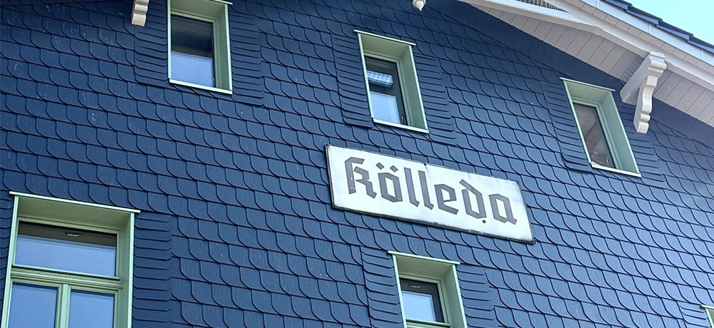 Aktuelles 2 Fassade des ehemaligen Bahnhofs Kölleda mit Schieferverkleidung und grün umrahmten Fenstern; mittig ist ein weißes Schild mit der Aufschrift „Kölleda“ angebracht.