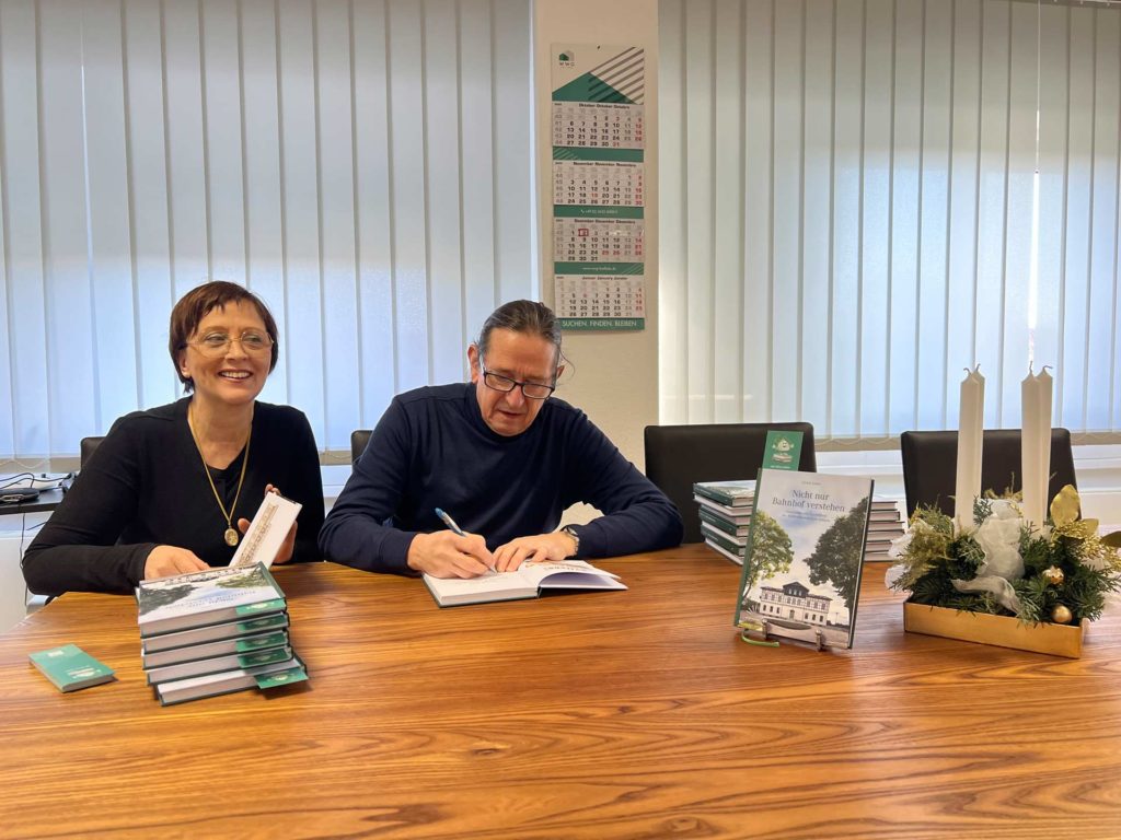 Christina Otto, Geschäftsführerin der WWG Kölleda, und der Autor Ulrich Seidel sitzen an einem Tisch und signieren Exemplare des Buches „Nicht nur Bahnhof verstehen“, das vor ihnen ausliegt.