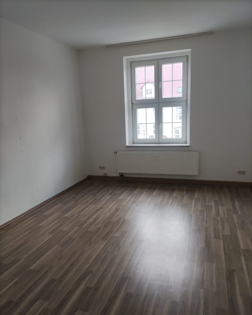 weiß gestrichenes Wohnzimmer mit dunklem Laminatboden, Fenster mittig mit Heizkörper direkt unter dem Fenster