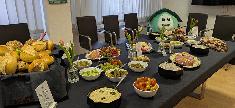 Aktuelles 2 Langer Konferenztisch mit dunkler Tischdecke, reich gedeckt mit Brötchen, Aufschnitt, Käse, Trauben, Tomaten, Dips, Gurken und weiteren Frühstücksbeilagen. Kleine Vasen mit Tulpen stehen auf dem Tisch. Im Hintergrund sind schwarze Stühle angeordnet; rechts hinten sitzt das WWG-Maskottchen Kölli.