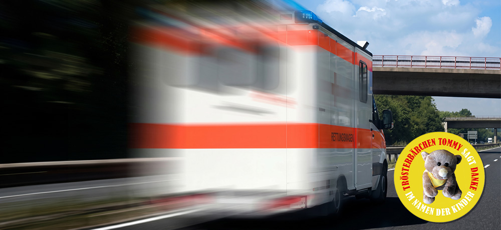 Schnell fahrender Rettungswagen auf einer Straße, aufgenommen mit starker Bewegungsunschärfe aus seitlicher Perspektive; im Hintergrund sind eine Brücke, Leitplanken und Bäume unter blauem Himmel zu sehen, rechts im Bild ein rundes Logo mit einem Teddybären und der Aufschrift „Trösterbärchen Tommy sagt Danke im Namen der Kinder“.