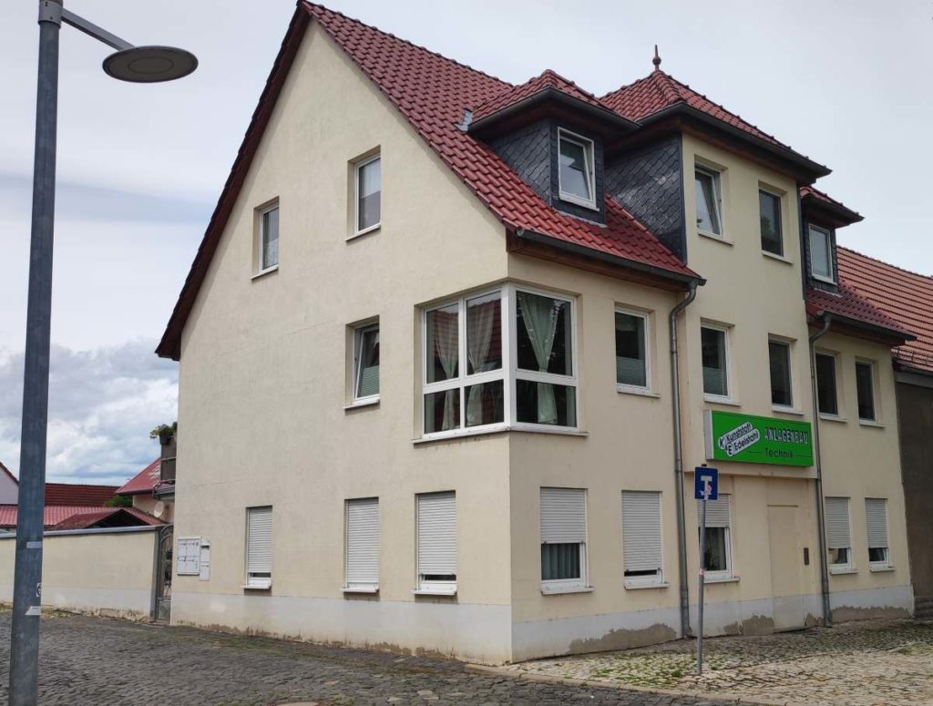 Das Bild zeigt ein mehrstöckiges, hell verputztes Gebäude m mit rotem Ziegeldach und Fenstergauben im Dachgeschoss, das an einer Straßenecke steht.